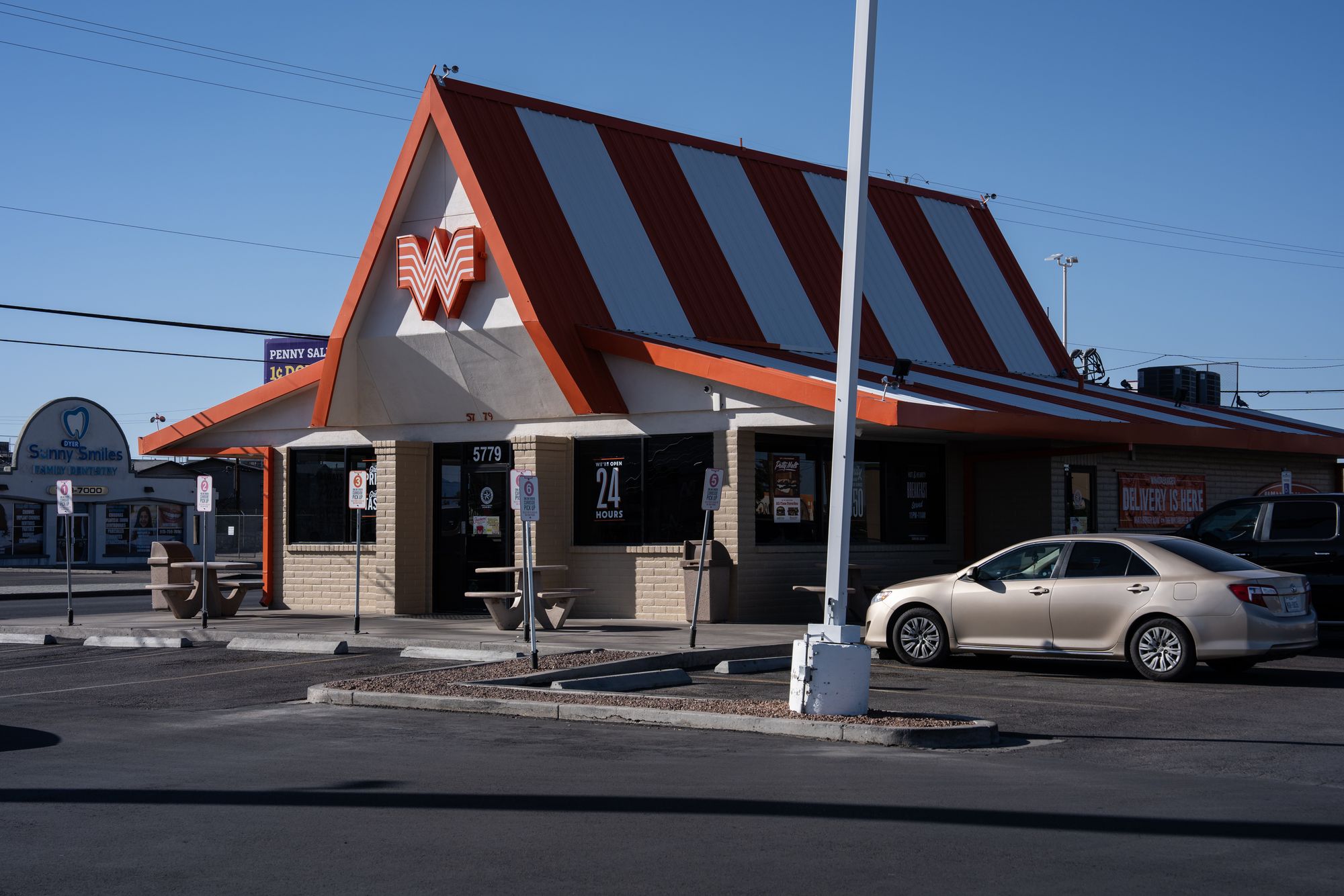 A photo shows a Whataburger fast-food restaurant. It has a triangular roof with orange and white stripes. The sky is blue above the restaurant, and there is a car parked outside and to the right. 