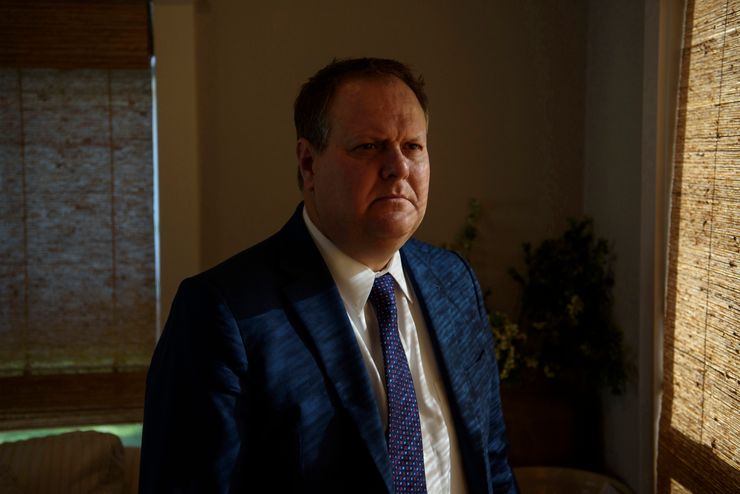 Chris Morton, a man with light-toned skin and wearing a blue suit jacket and tie, stands near a window.  