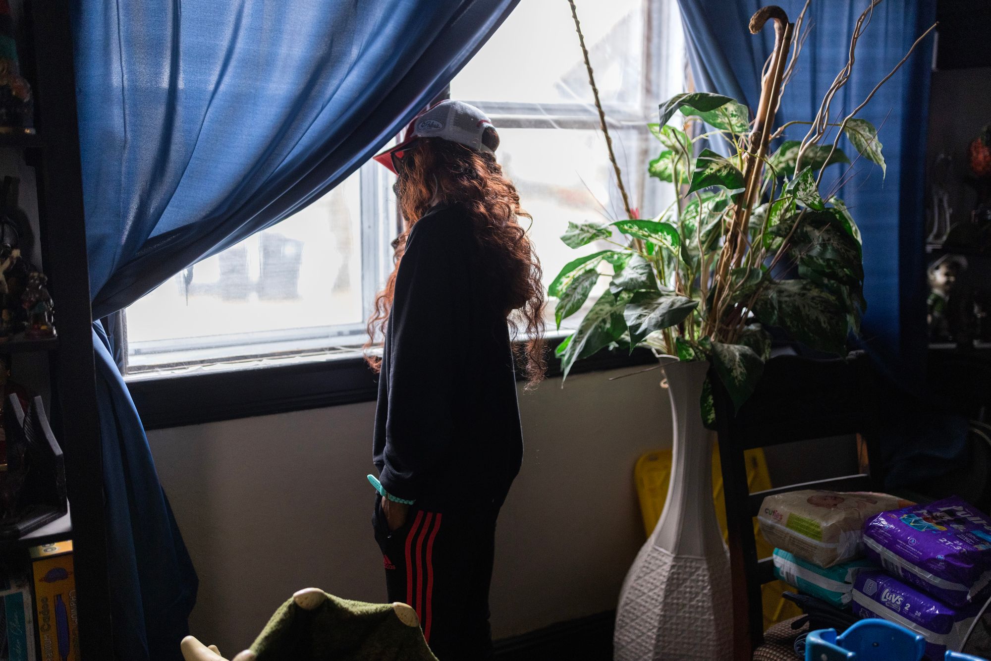 A woman with long, wavy red hair, whose face is not visible, looks out a window with blue curtains. Behind her is a tall vase with green leaves in it. A stack of diapers is on a chair.  