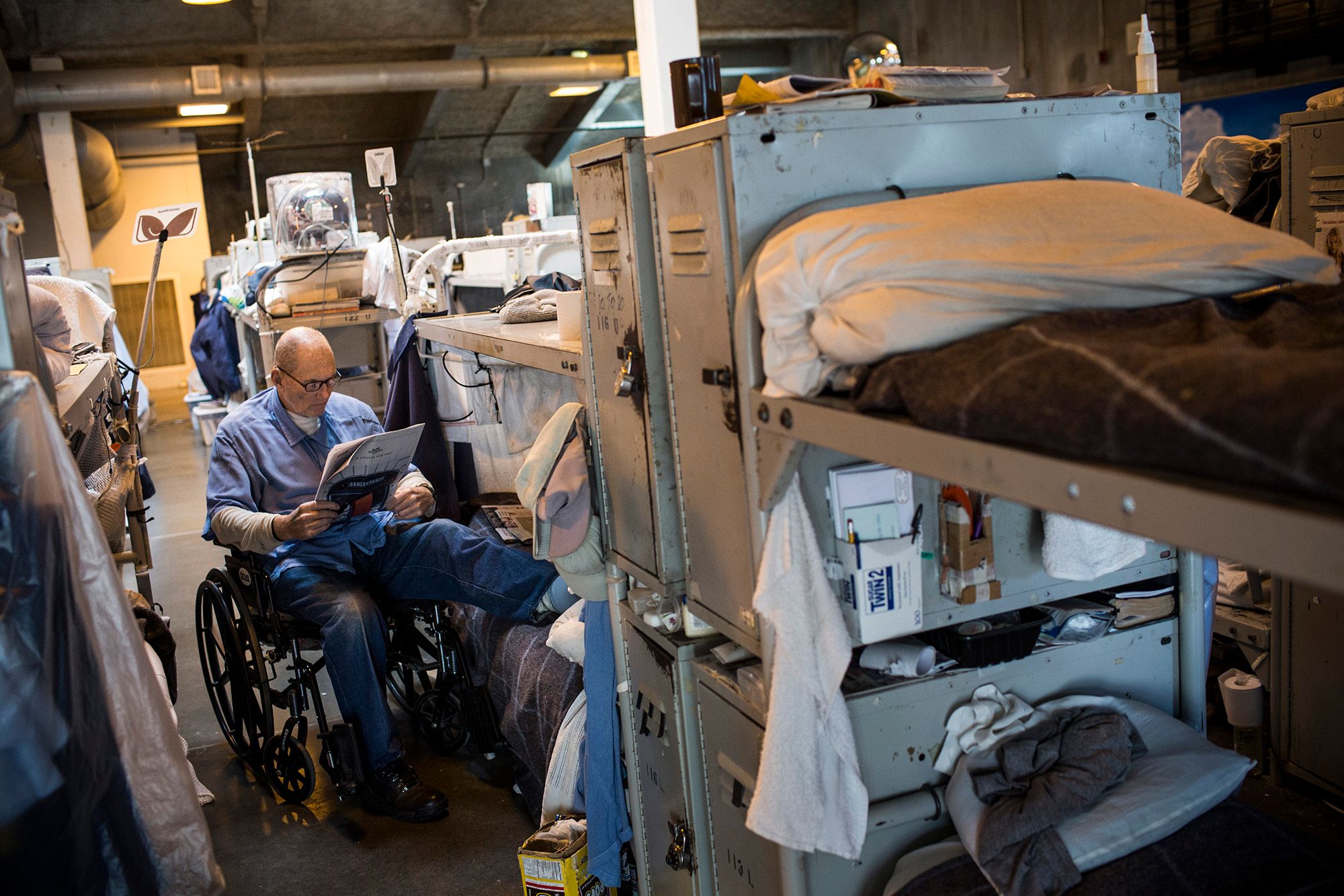 An elderly man, wearing glasses and a blue prison uniform, is reading a newspaper. He sits in his wheelchair, which is next to a couple of bunk beds within his prison cell block.