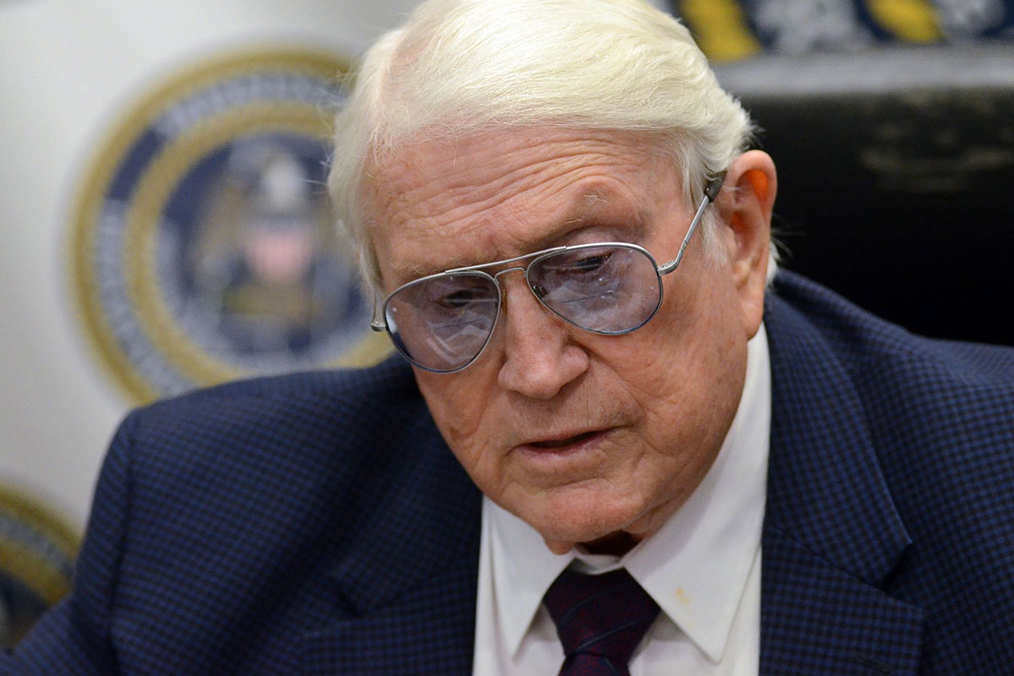 A close-up photo of an older White man with white hair, blue-tinted glasses, and a navy blue suit. 