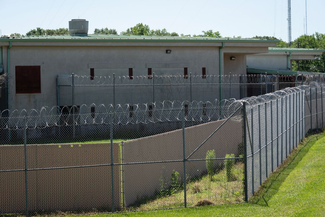 A photo shows a beige building with a sea-green roof. Barbed wire fencing surrounds the building. 