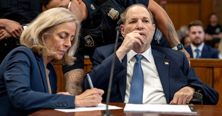 A White man, wearing a navy blue suit, is handcuffed to a chair as he sits next to his attorney in a courtroom. Two court officers stand behind them. 