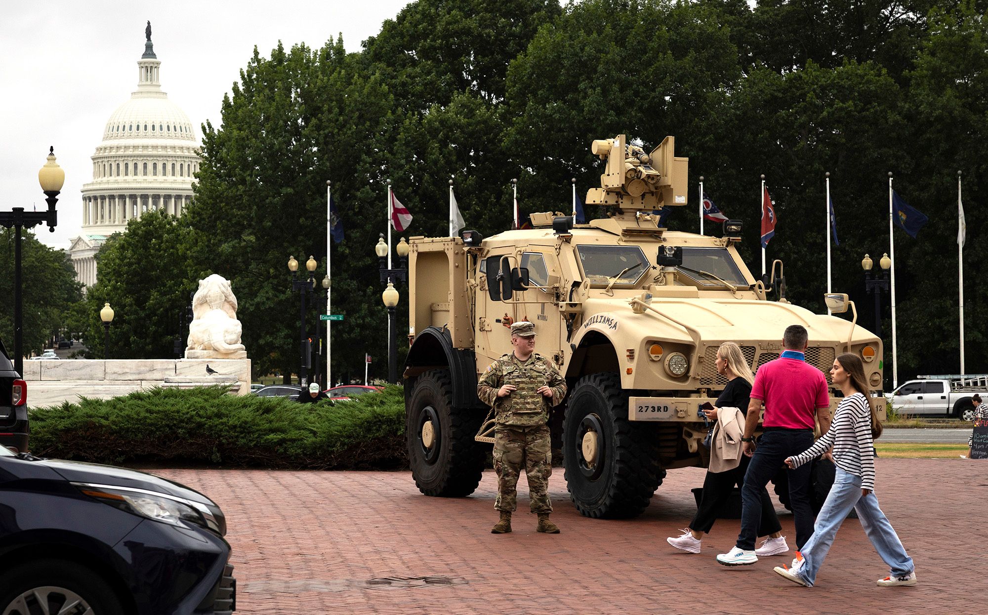 A person in a National Guard uniform stands next to a tan military vehicle parked on a red brick pavement. Three people walk in front of the military vehicle. The U.S. Capitol Dome and various trees can be seen in the background. 
