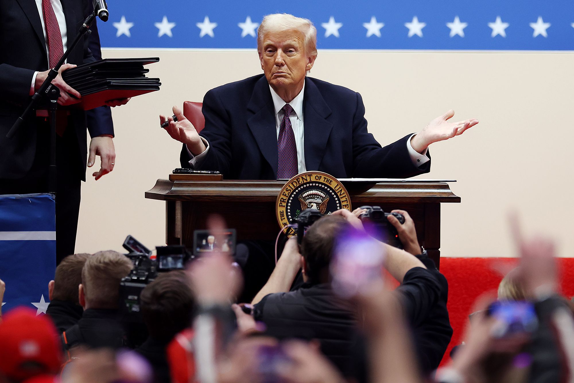 President Donald Trump, a White man wearing a dark suit, gestures with his hands out while sitting at a desk with a presidential seal on it. On the left, a man holds a stack of executive orders.  In front of the president, photographers take pictures of him. 