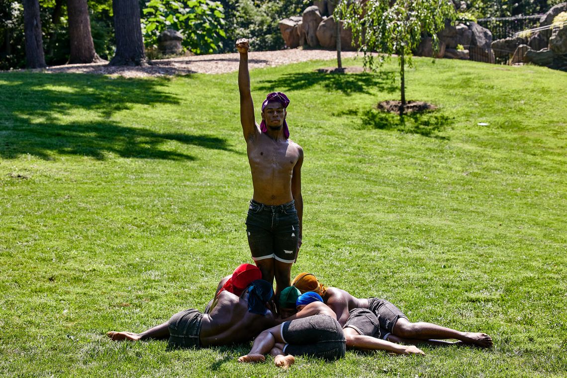 Khayr Muhammad, wearing a purple velvet durag and with his arm raised in a Black Power salute, stands in the center of a field of green grass, surrounded with dancers lying down near his feet. 