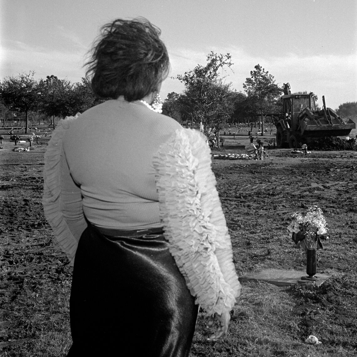 A Black woman in a blouse with ruffled sleeves stands with her back to the camera facing a gravestone that has a vase of flowers set upon it.
