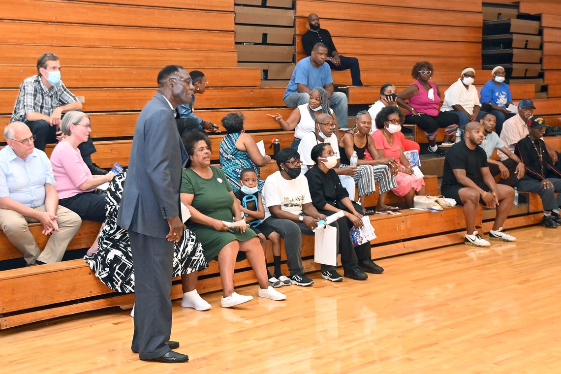 A Black man wearing a gray suit stands while speaking in a gymnasium as a group of residents sits on the bleachers. 