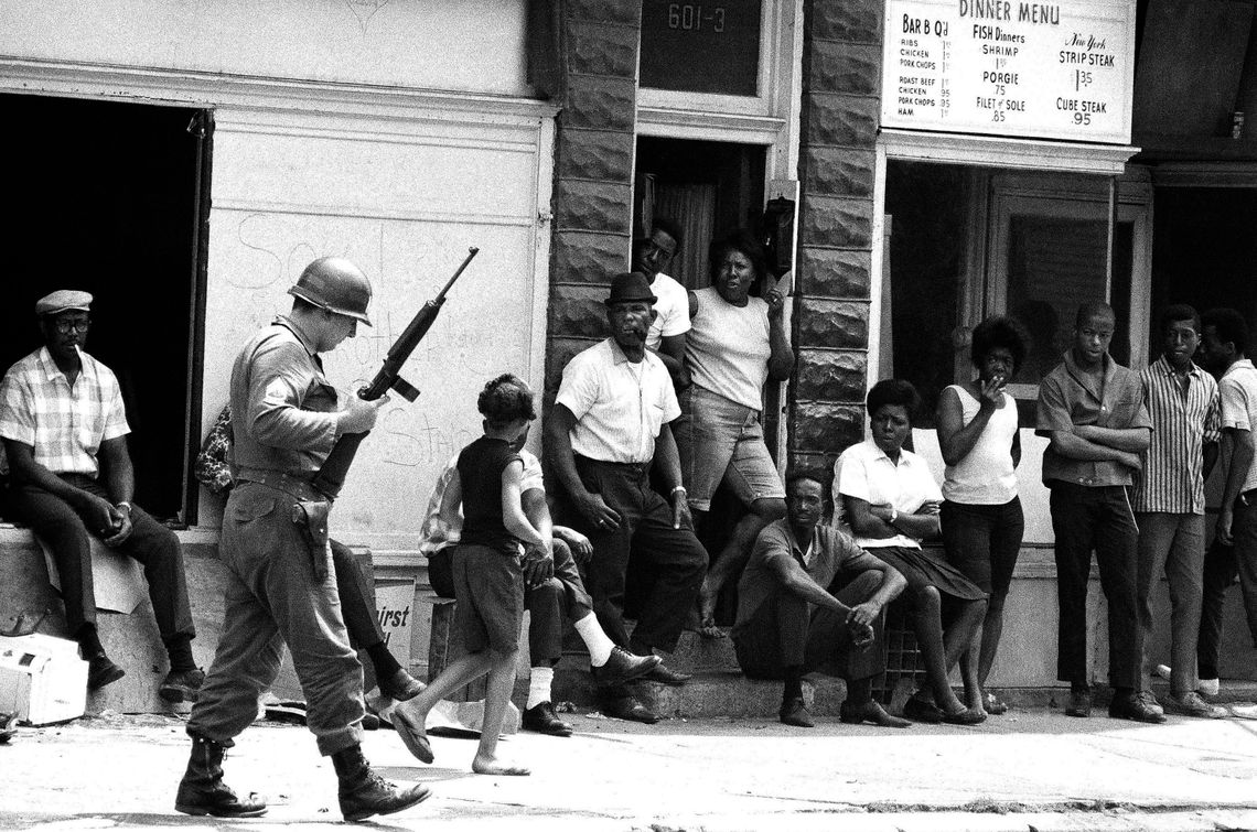 A black-and-white photo shows a National Guard member with a rifle and helmet walking down a street past a line of Black men and women standing against the buildings watching him. 