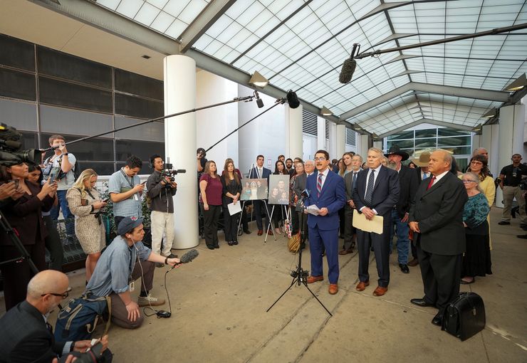 Travis County District Attorney José Garza, a Mexican man in a blue suit, speaks to reporters while a group of people stand behind him with portraits on printed boards.  