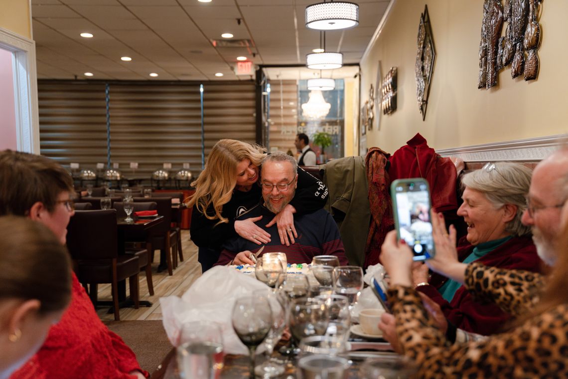 A photo shows Sandra, a white woman, hugging a seated Latif, a Moroccan man, at a restaurant. Five people are sitting around them at the table, and one woman with light-toned skin is holding her phone up to take a picture. 