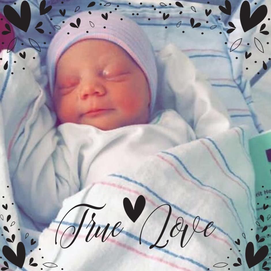 A photo of a sleeping newborn in a white, pink and teal blanket. Cursive black text on top of the image reads “True Love” with a black heart between the words. 