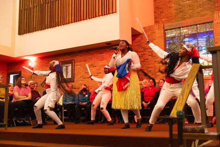 A photo shows a Black woman wearing a grass skirt and holding a blue-and-red Haitian flag while singing into a microphone. Dancers holding toy swords surround her as they all perform on a church stage.