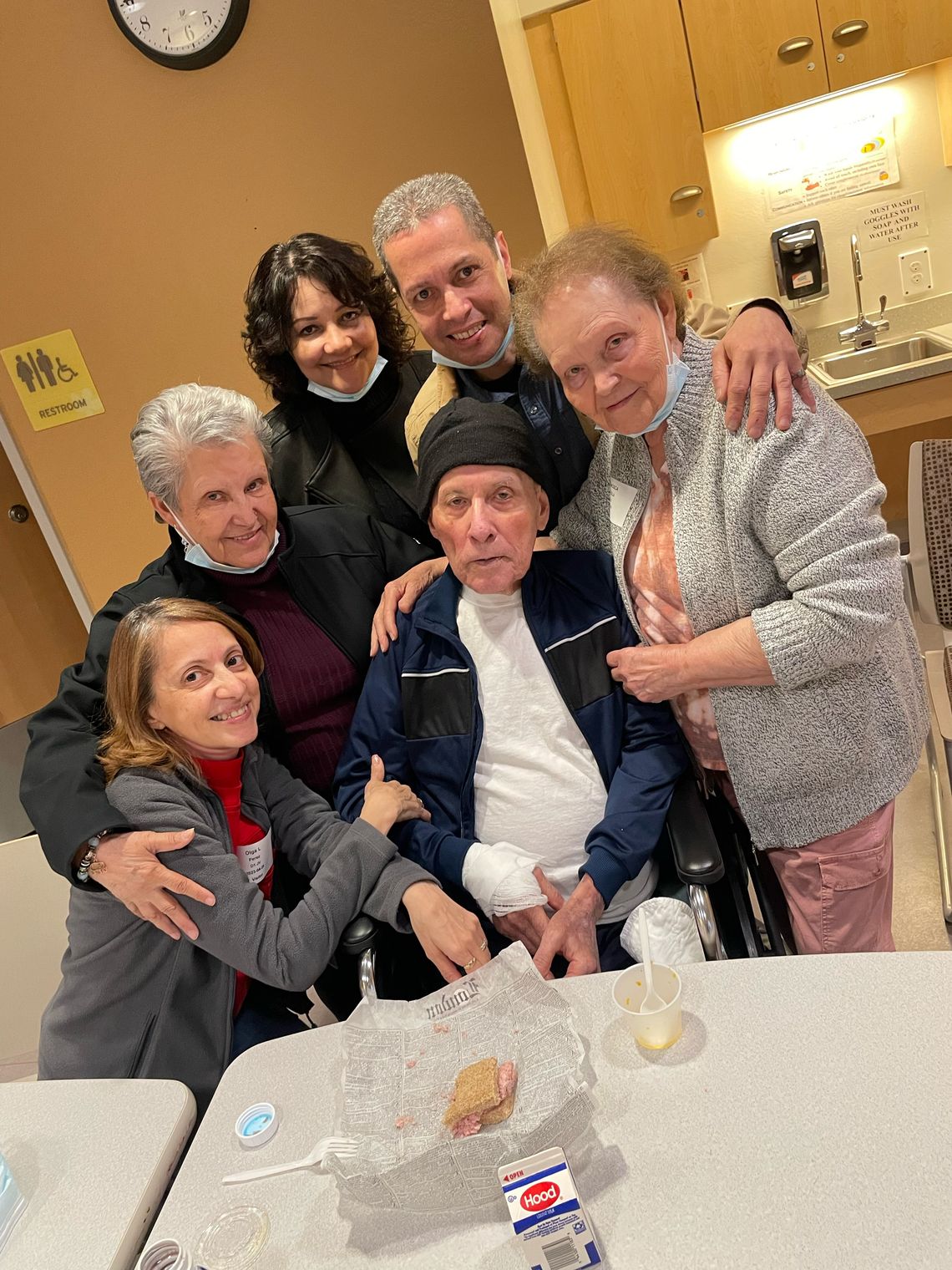 An older man sitting near a table in a hospital facility wears a black beanie and a black and blue jacket. His daughter, sisters, granddaughter and son surround him to pose for a photo.