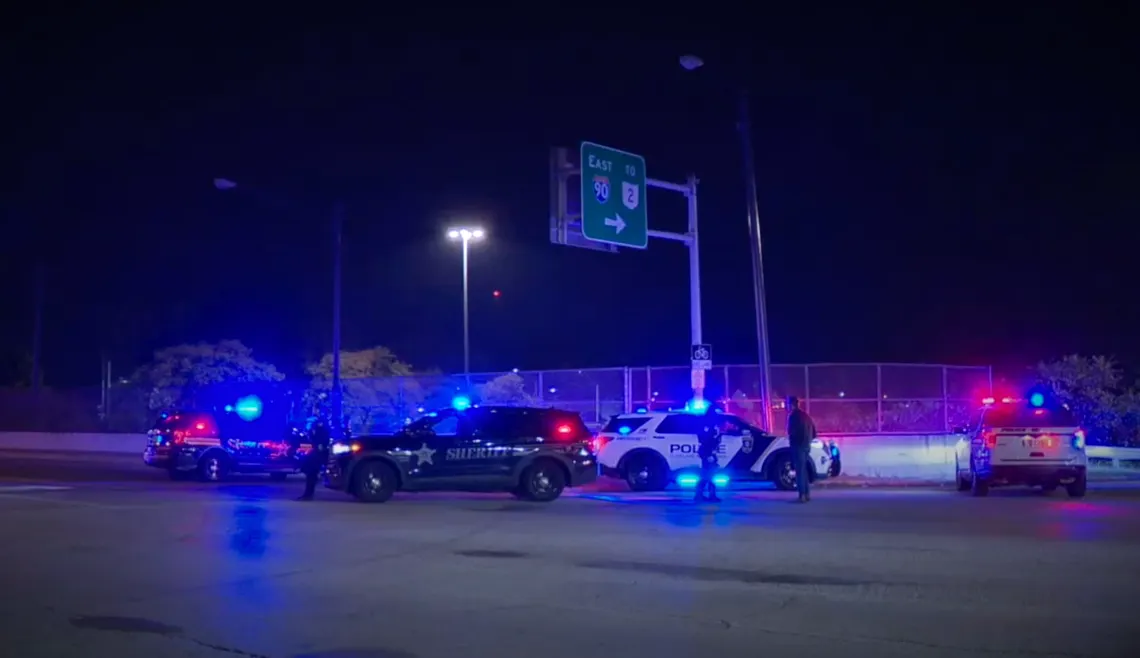 Four law enforcement vehicles are parked near each other, with their red and blue lights on at night, under a highway sign that points toward I-90 and Route 2.