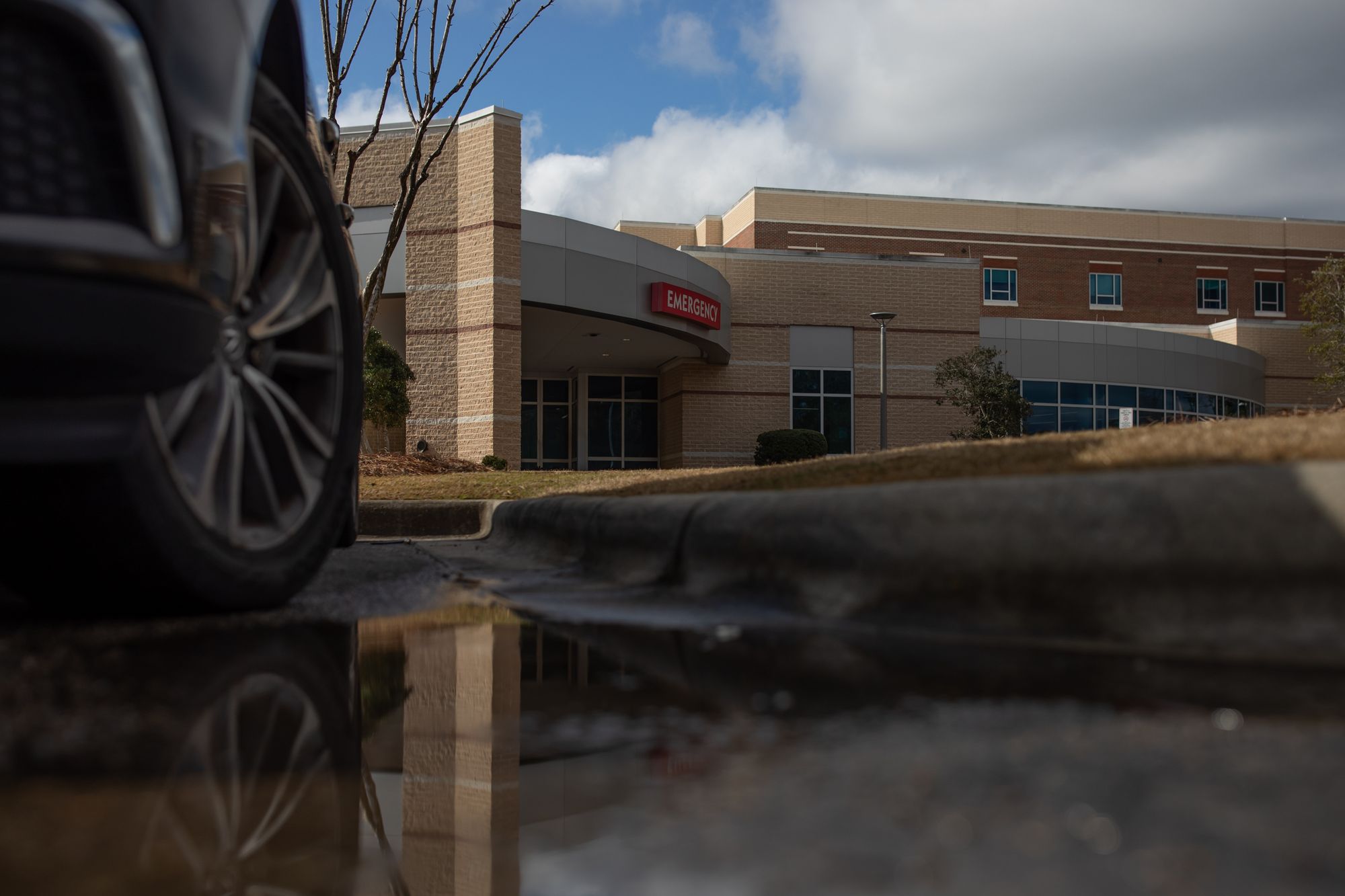 A photo shows a hospital’s emergency entrance. In the foreground, a car’s passenger-side wheel is visible.