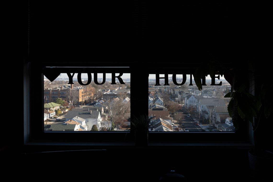 A photo shows the silhouette of a banner that reads “YOUR HOME”, hanging from the top of a window frame. The city of Bayonne is visible through the window. 