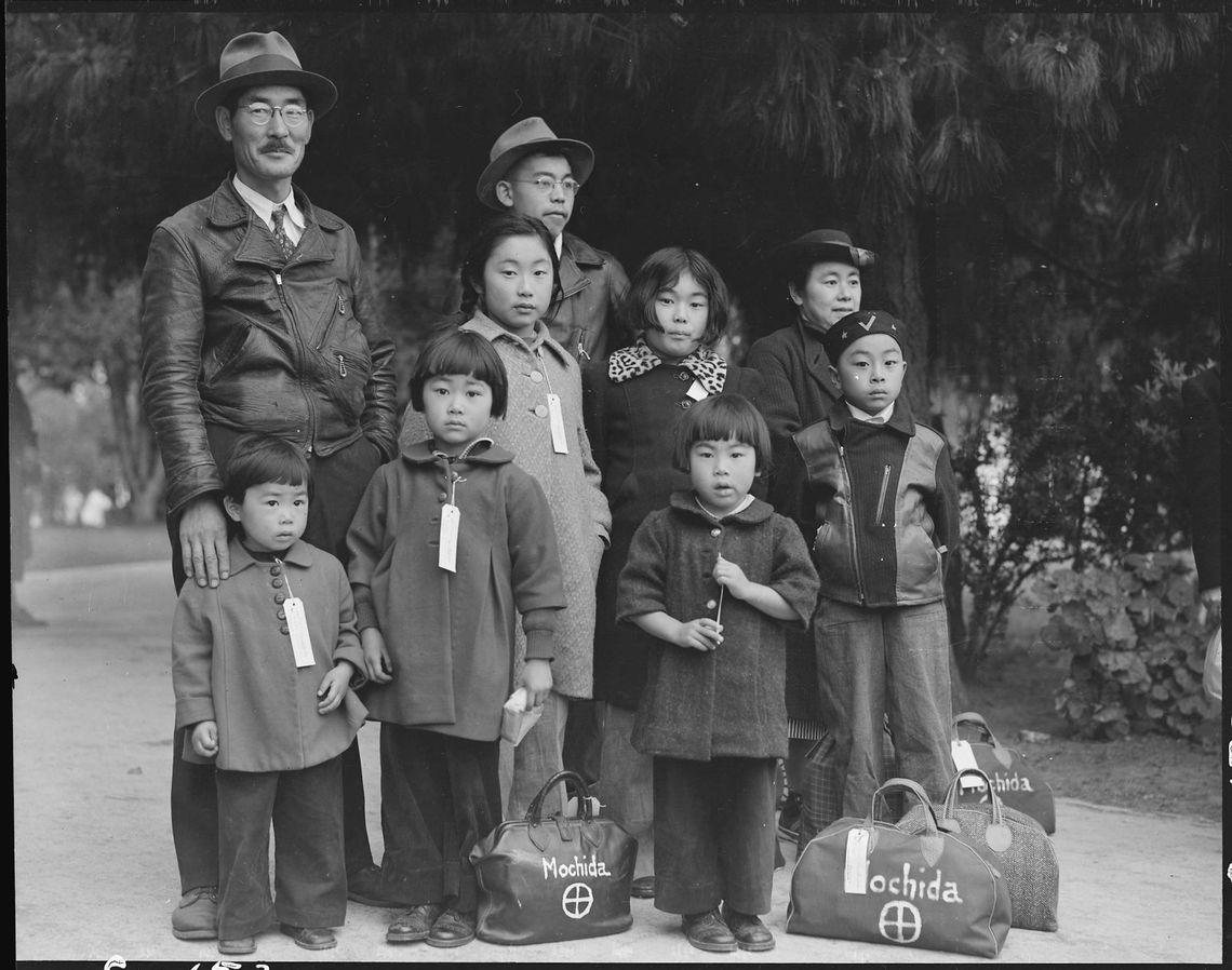 A black-and-white photo shows a family of nine, dressed in coats and standing outdoors. Some of the children have paper tags hanging from their coats. 