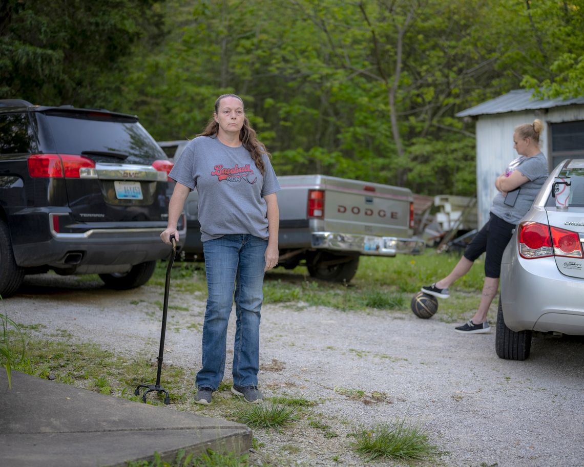 Helphenstine, a White woman wearing jeans and a gray T-shirt and holding a walking stick, stands in front of two trucks. Another White woman leans against a car on the right side of the photo.