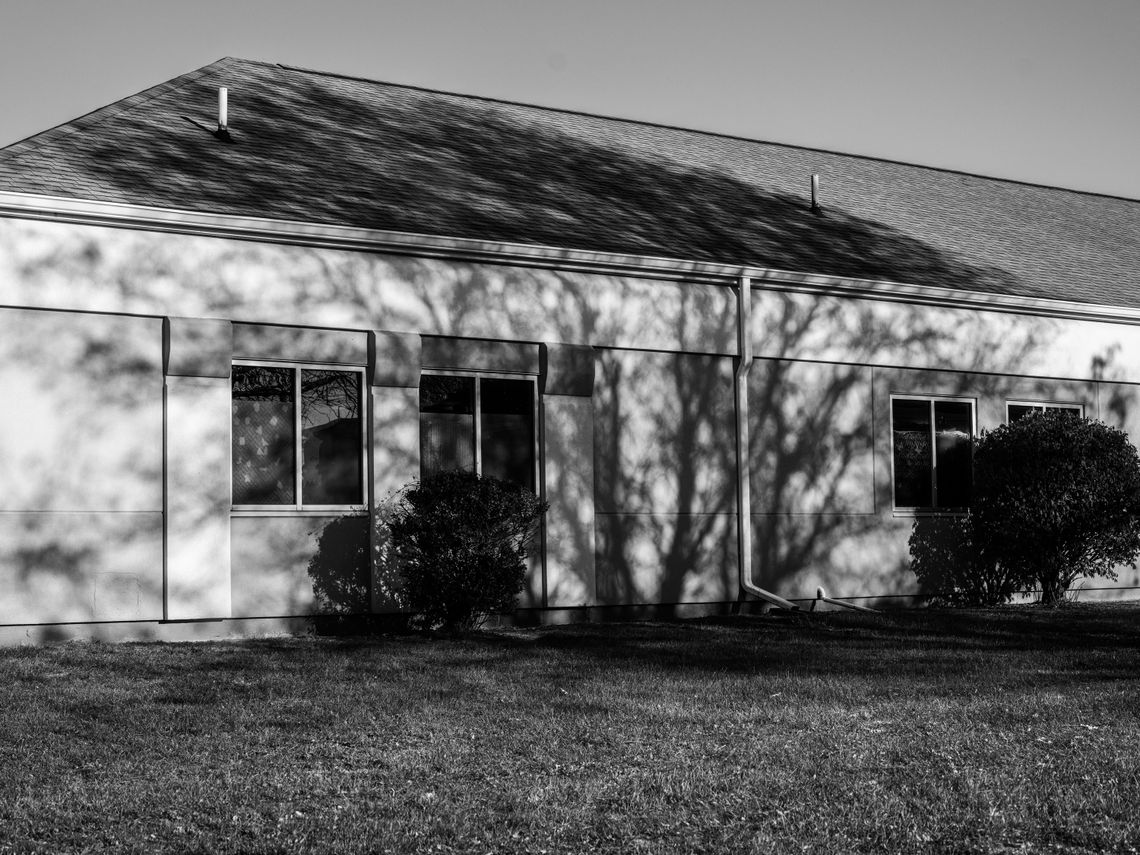 A black-and-white photo shows a one-story building with a grey roof. The shadows of trees are visible on the side of the building.