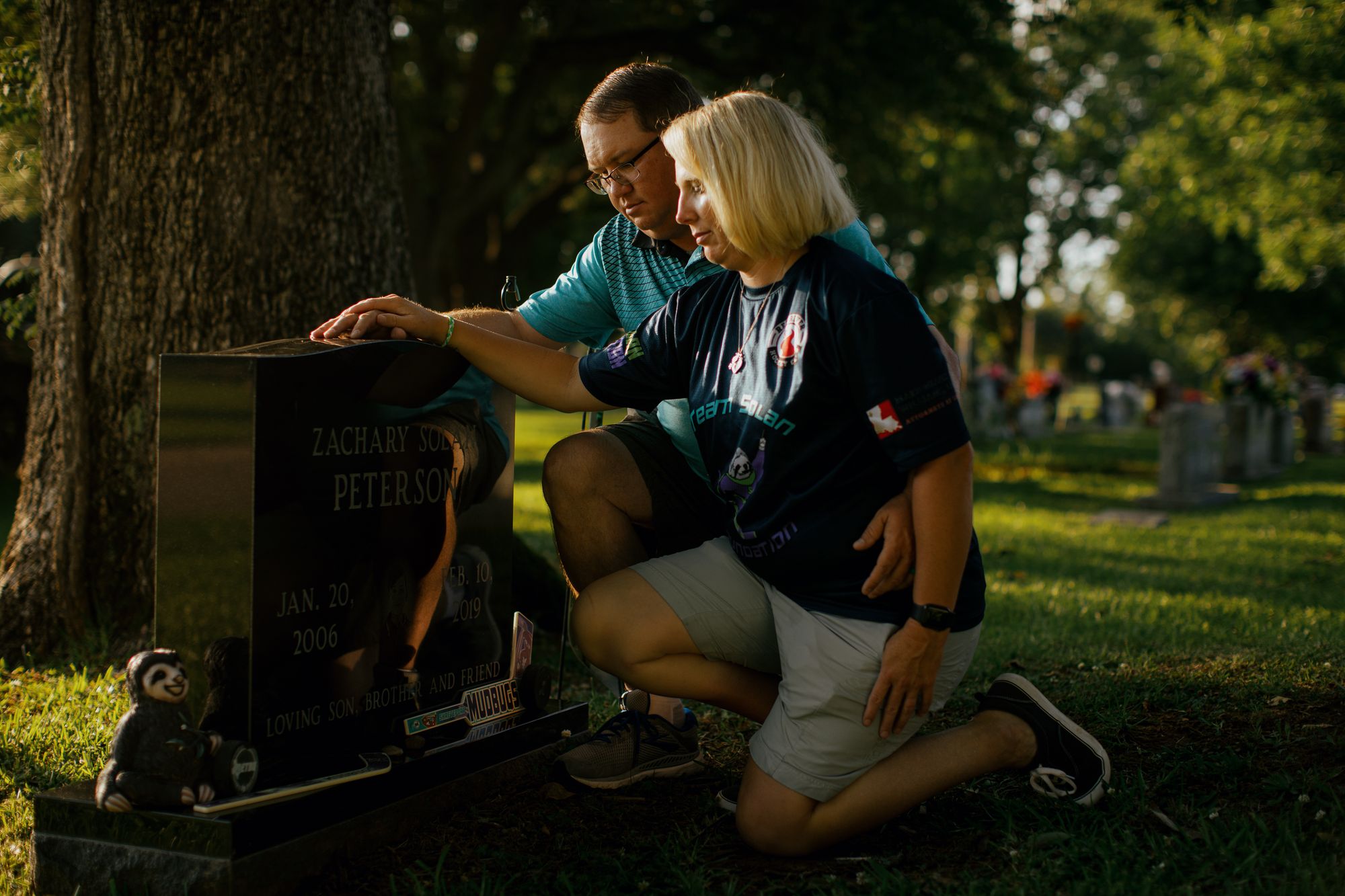 Ronnie Peterson, a man with short brown hair, light-toned skin and wearing a turquoise polo shirt, and Bridget Peterson, a woman with medium-length blonde hair, light-toned skin and wearing a navy blue T-shirt, kneel at the grave of their son, with their hands resting on the gravestone.  