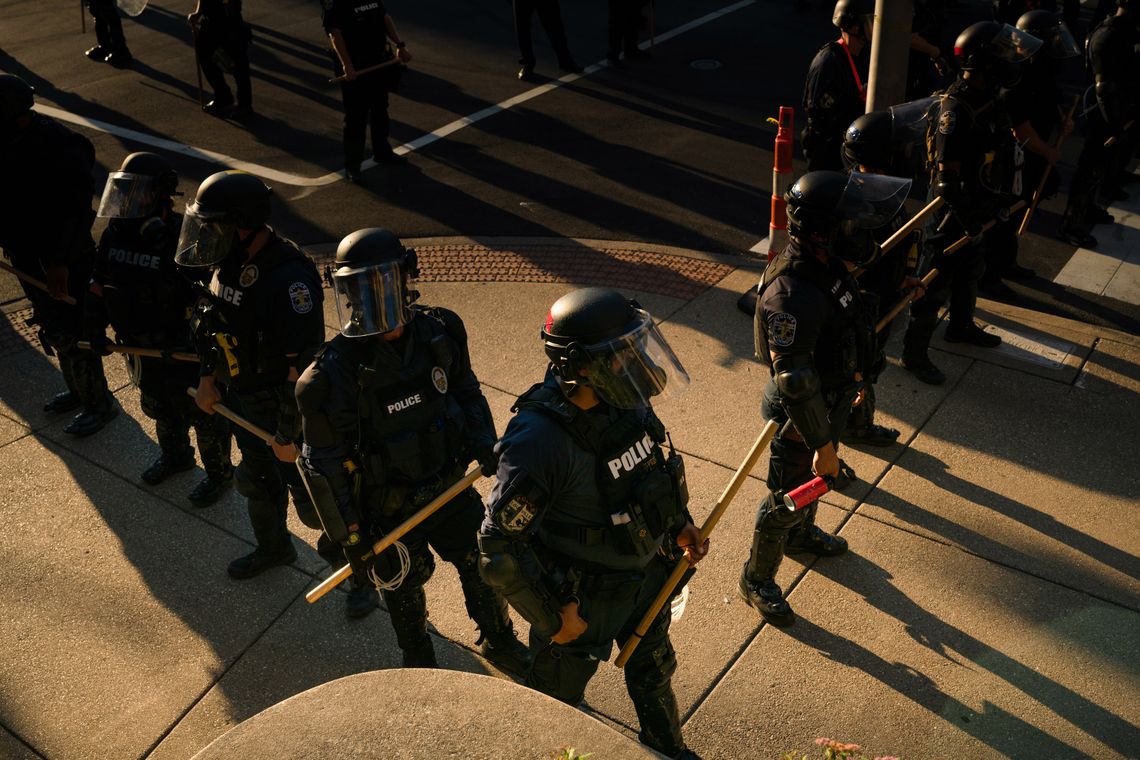A photo shows a line of police officers standing on a corner while wearing riot gear and holding batons. 