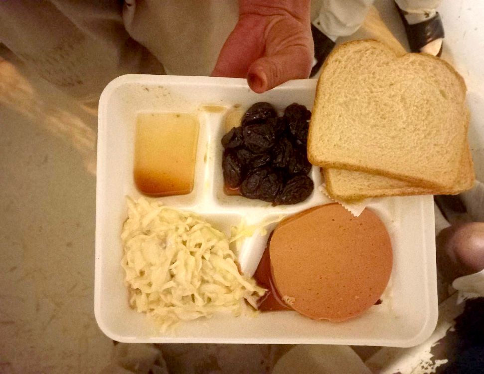 A white tray with coleslaw, prunes, two pieces of bread and baloney. 