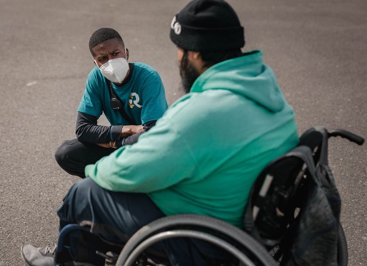 A Black man, wearing a KN95 mask, black long-sleeved undershirt and teal T-shirt, kneels down in front of a bearded man sitting in a wheelchair. The man in the wheelchair is wearing a black beanie, a seafoam green sweater and jeans. A gray backpack hangs from the back of the wheelchair. 