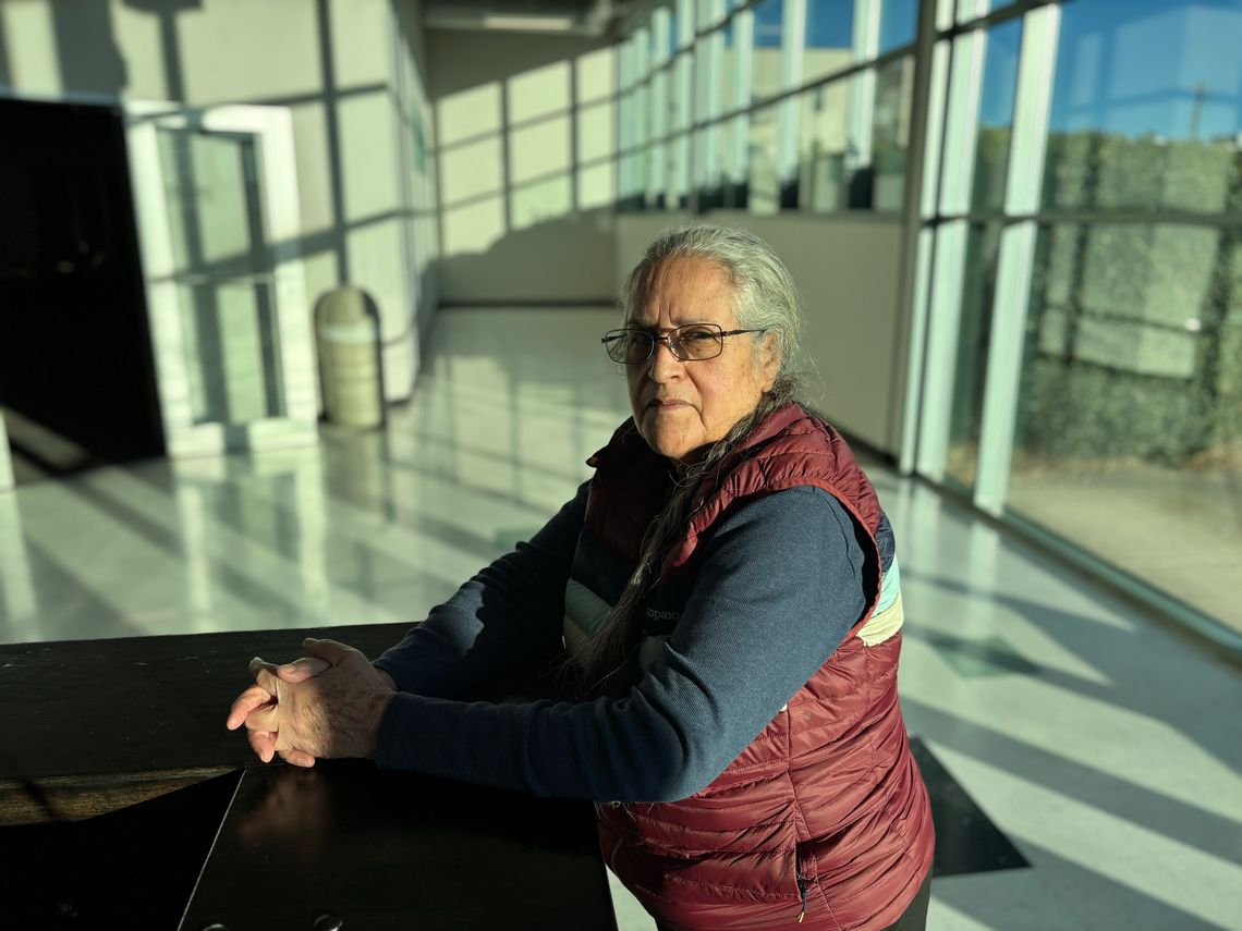 Margarita Solorzano, a Hispanic woman with white hair and wearing a maroon-colored vest and dark blue long-sleeved shirt, sits in a space near a wall of windows as light streams in. 