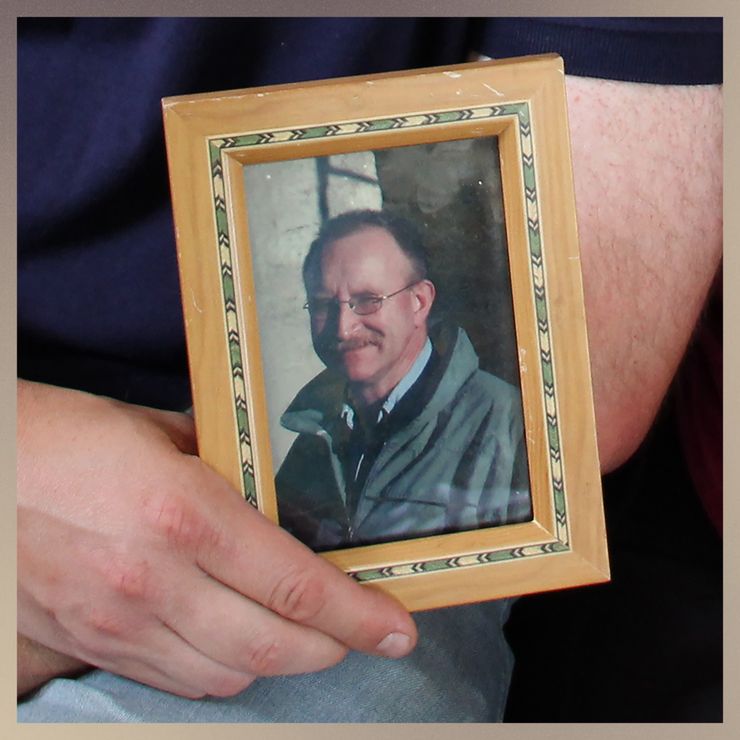 A person’s hand holds a framed photo of a White man with a mustache, who is wearing glasses, a green jacket and a light blue button-down shirt. 