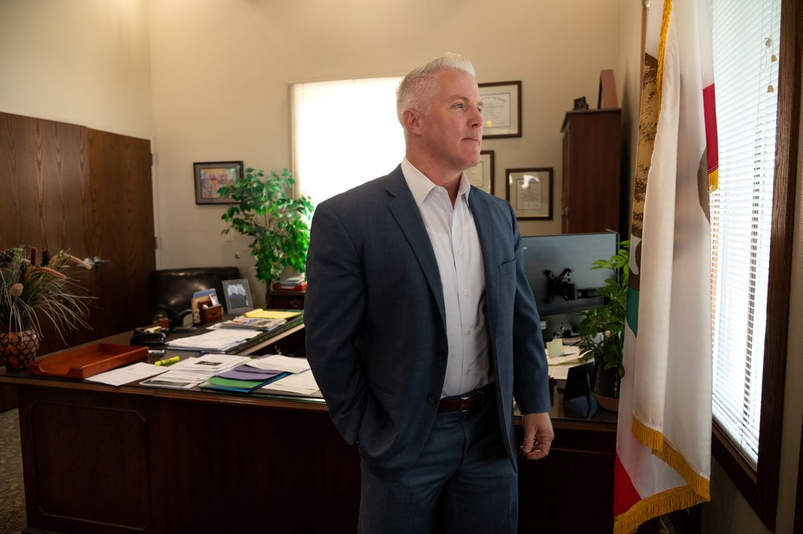 A White man with short white hair, wearing a suit, stands in his office. 