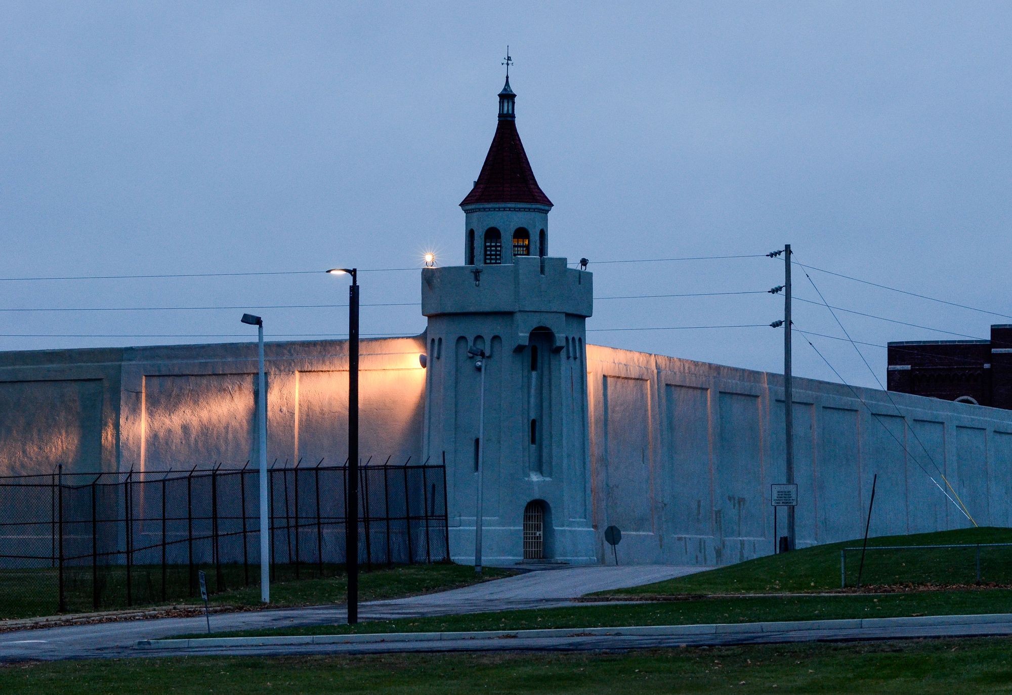 A photo of Attica at dusk shows high walls and a tall tower. The light falling on the walls is blue toned, reflecting the sky, and two lights beam out orange light from the tower. 
