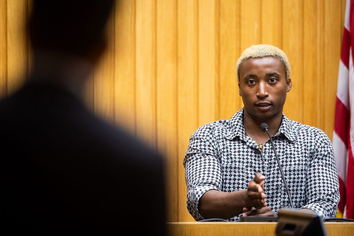 A Black person speaks into a courtroom microphone while sitting at a witness stand. 