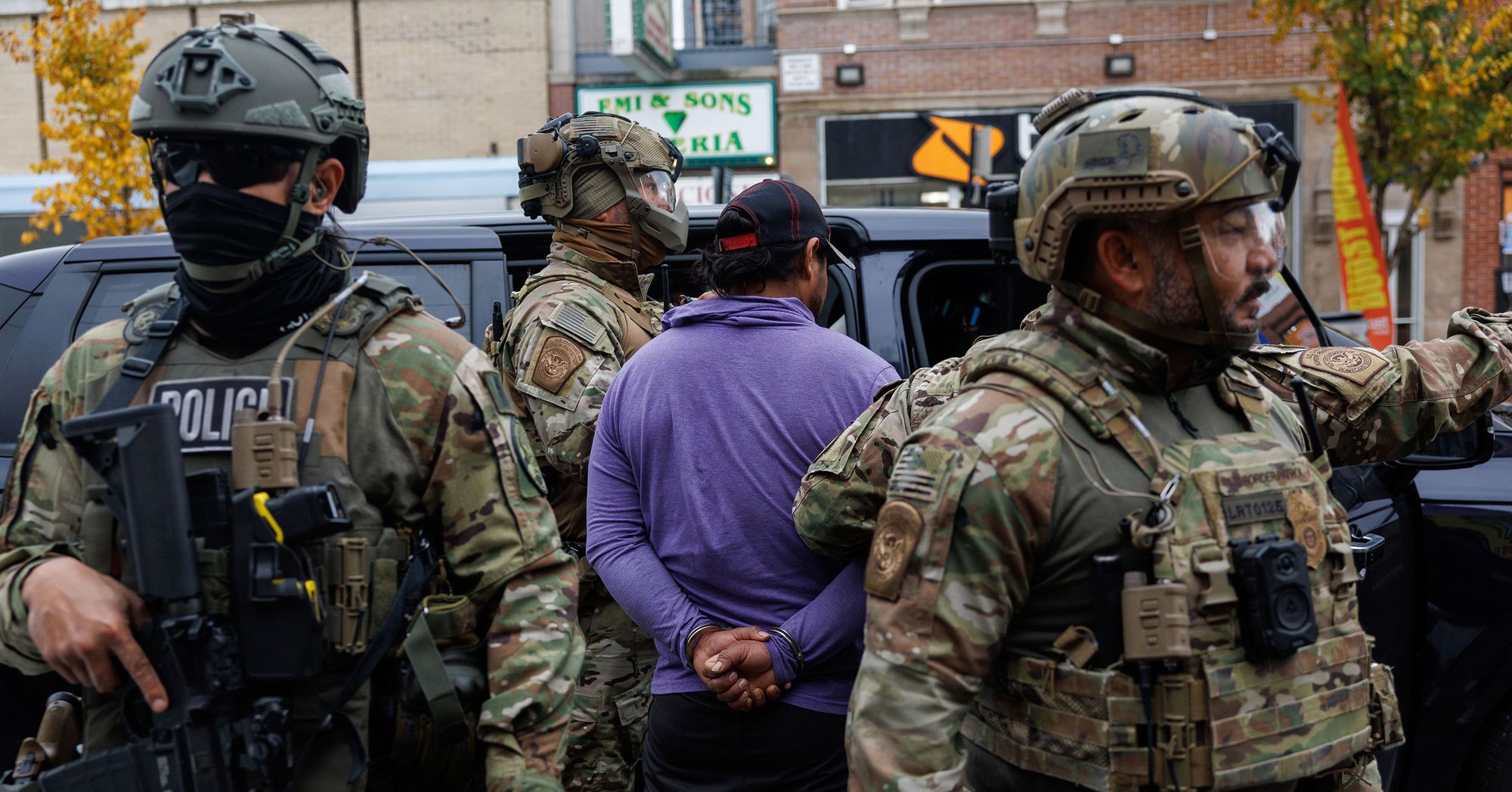 A man in a purple shirt has his hands cuffed behind his back while facing an open car door. Four federal officers, wearing camouflage uniforms, vests, and helmets, stand around the man. 