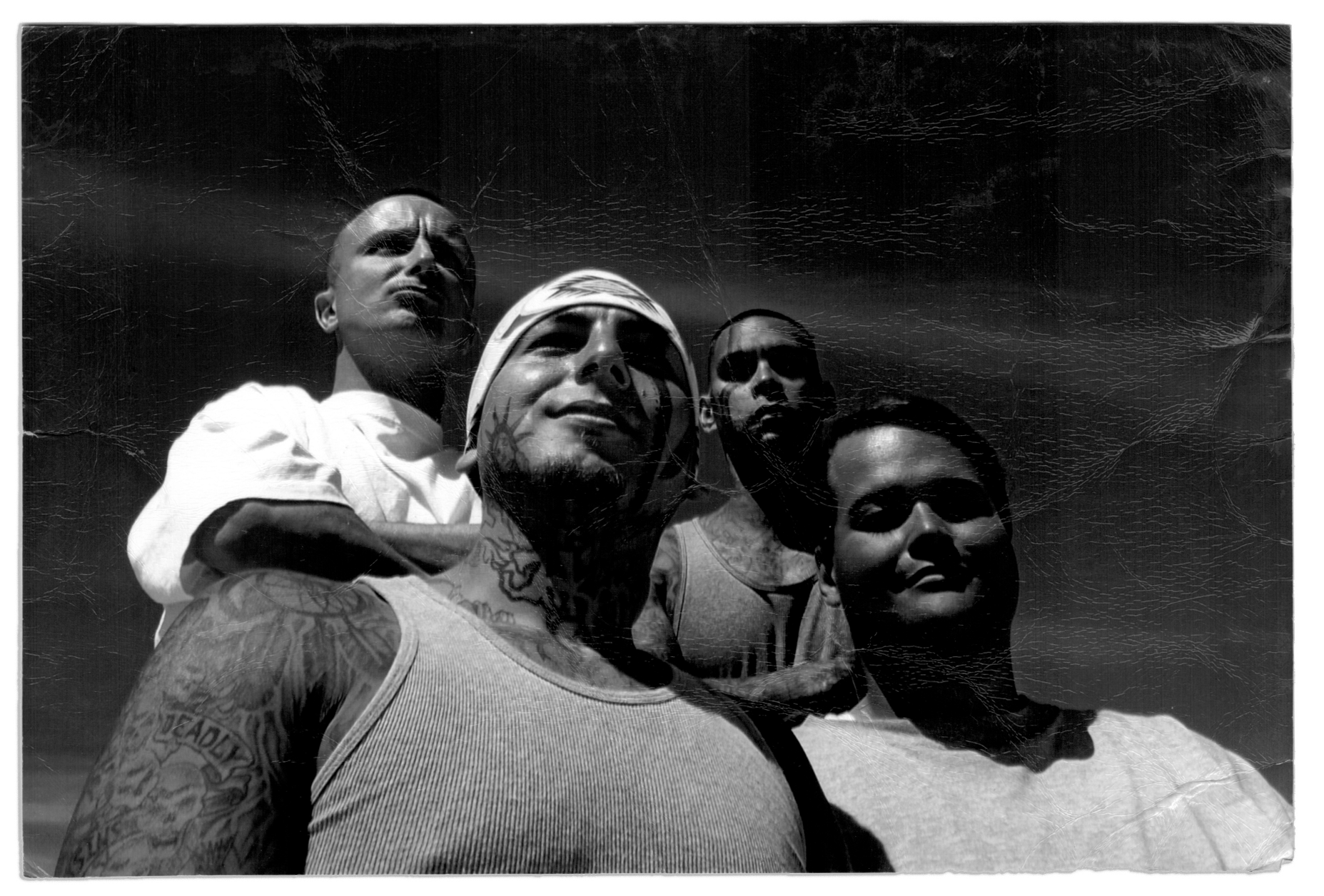 In a black and white photograph, Grace Pinson, a trans Latina with short dark hair, stands outside in the sun with three men. The person to her left wears a tank top and has tattoos on his face, neck and arms. Behind her stand two men. The one on the right wears a white T-shirt and is bald. The one on the left has tattoos on his chest and arms and has close-cropped, dark hair. 