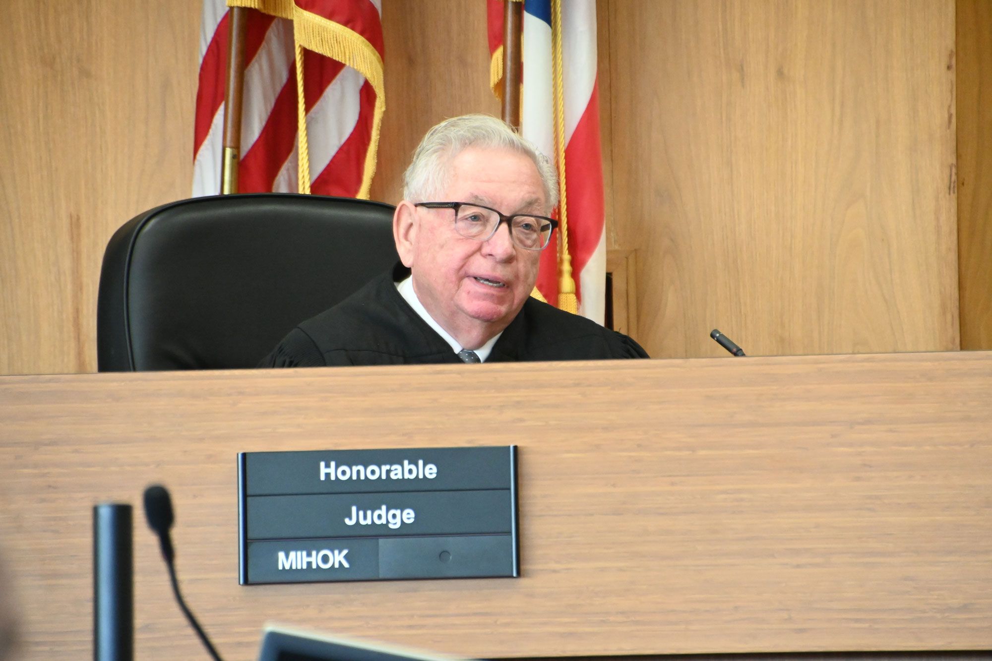 A White man with white hair wearing dark-rimmed glasses and a black judge’s robe sits in a large black chair at the bench, with the American and Ohio flags behind him.