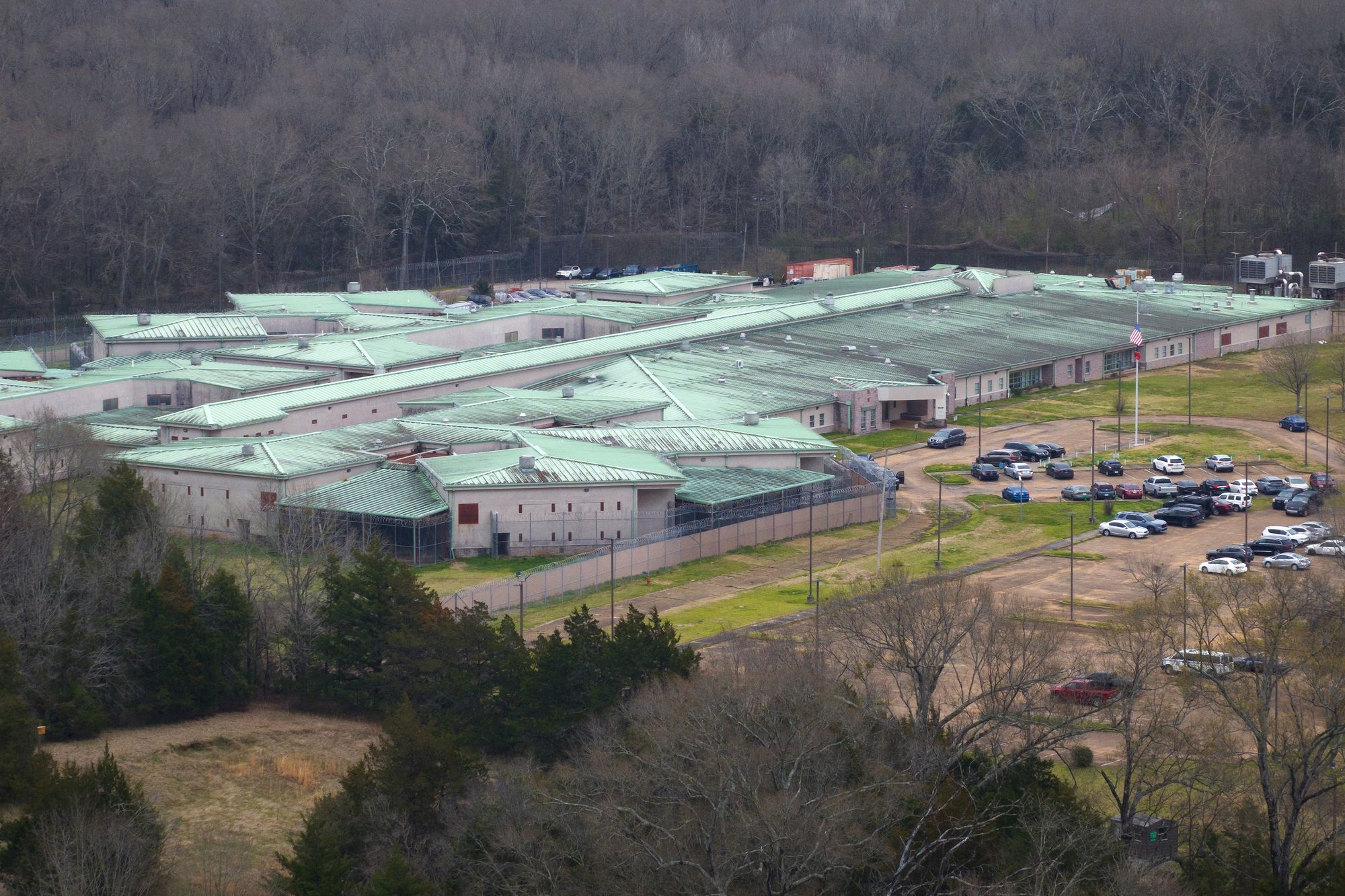 An aerial photo of a prison facility with a light teal roof and its adjacent parking lot. Bare trees surround the facility. 