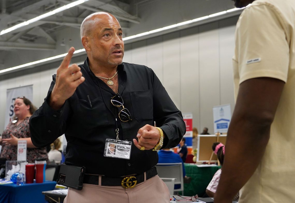 A Black man in a black button-down shirt gestures while speaking with a person wearing a light yellow shirt at a resource fair. 