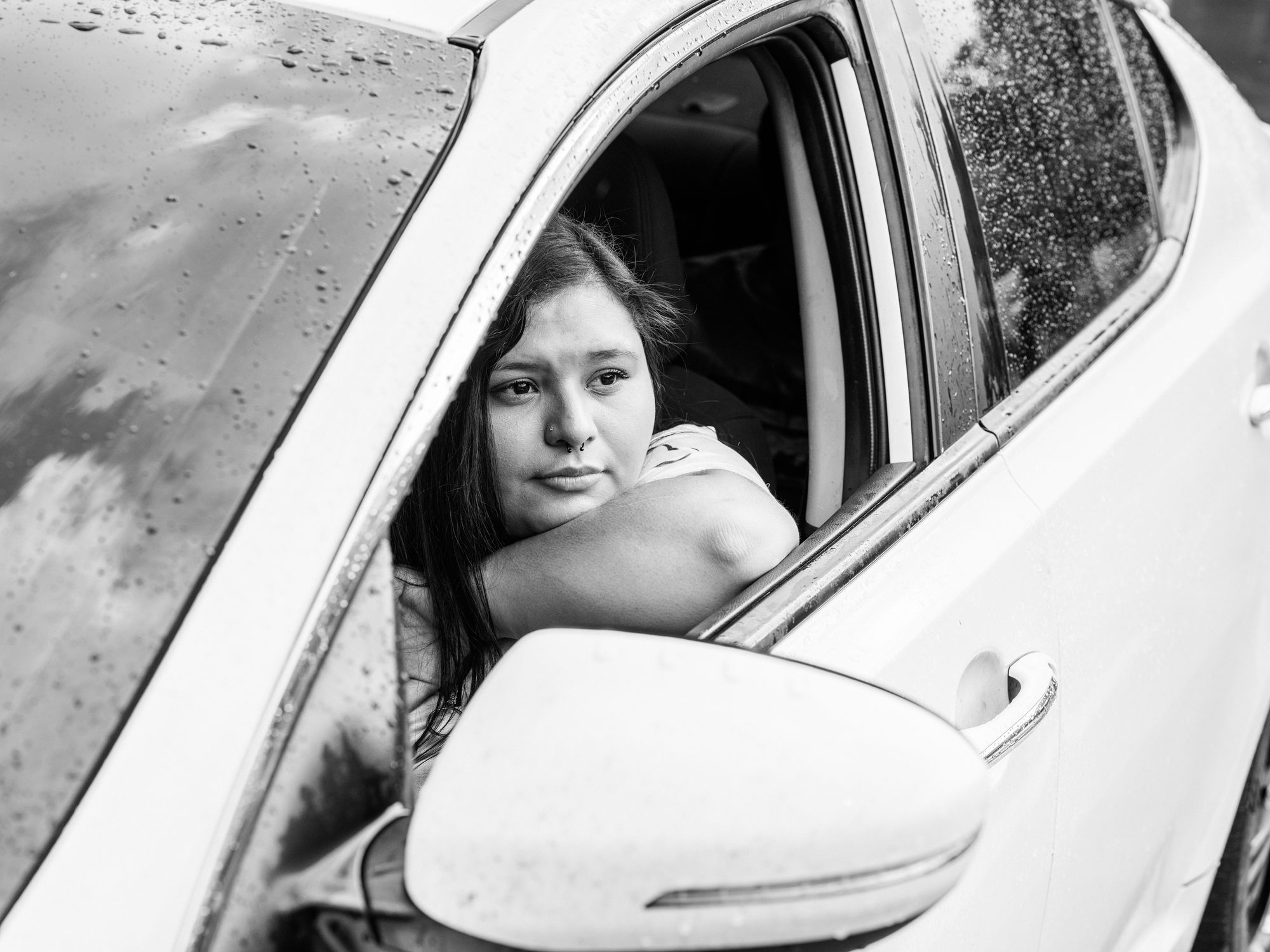 A black-and-white photo shows a Mexican American woman sitting in the driver’s seat of a car, looking out the window. 