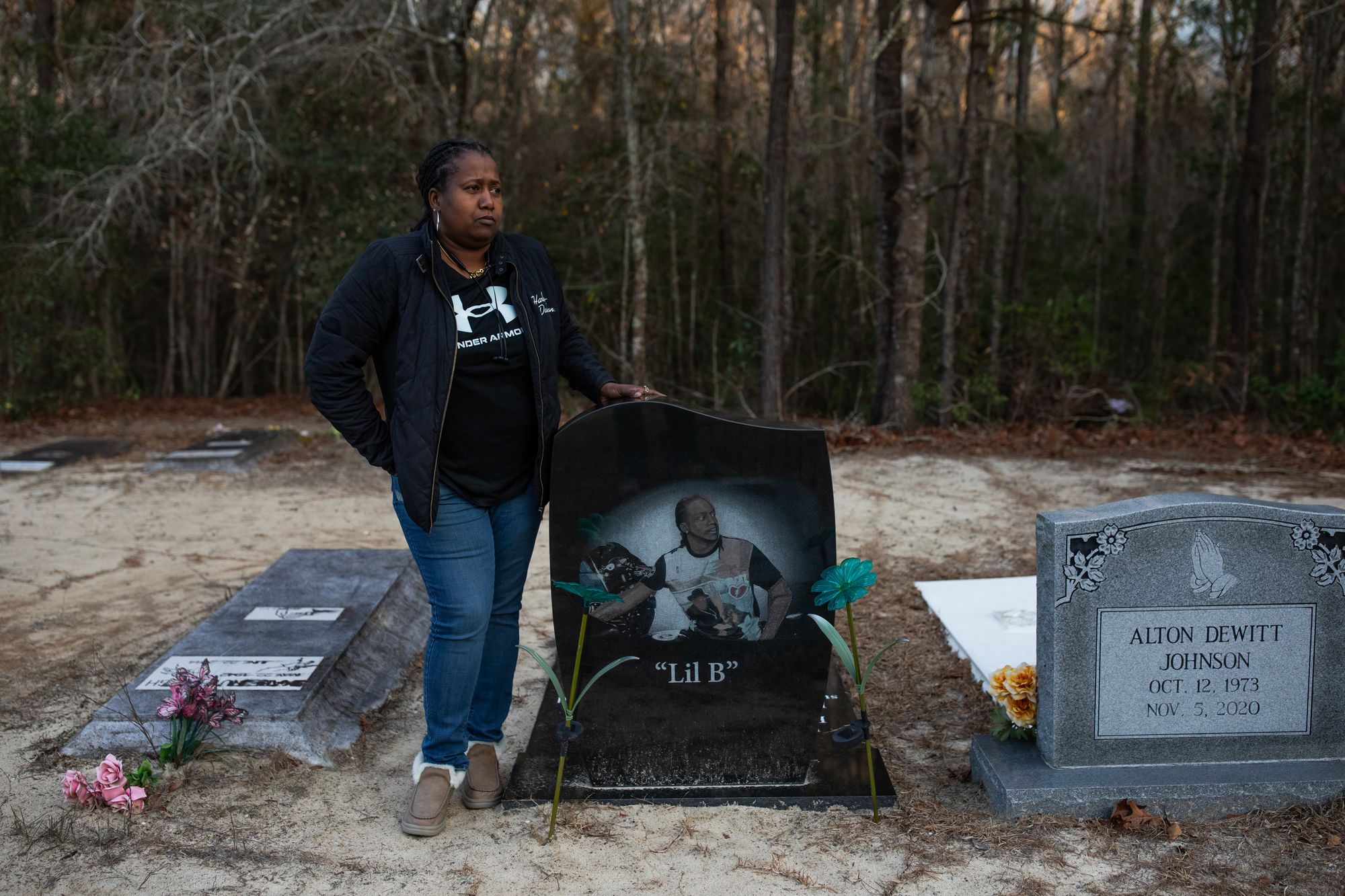 A photo shows a Black woman wearing a black jacket, T-shirt and blue jeans standing next to a gravestone with a picture of her son, a Black man, on it. The gravestone also has “Lil B” written on it. A few other graves are visible near her son’s gravestone, and trees are visible behind her.    