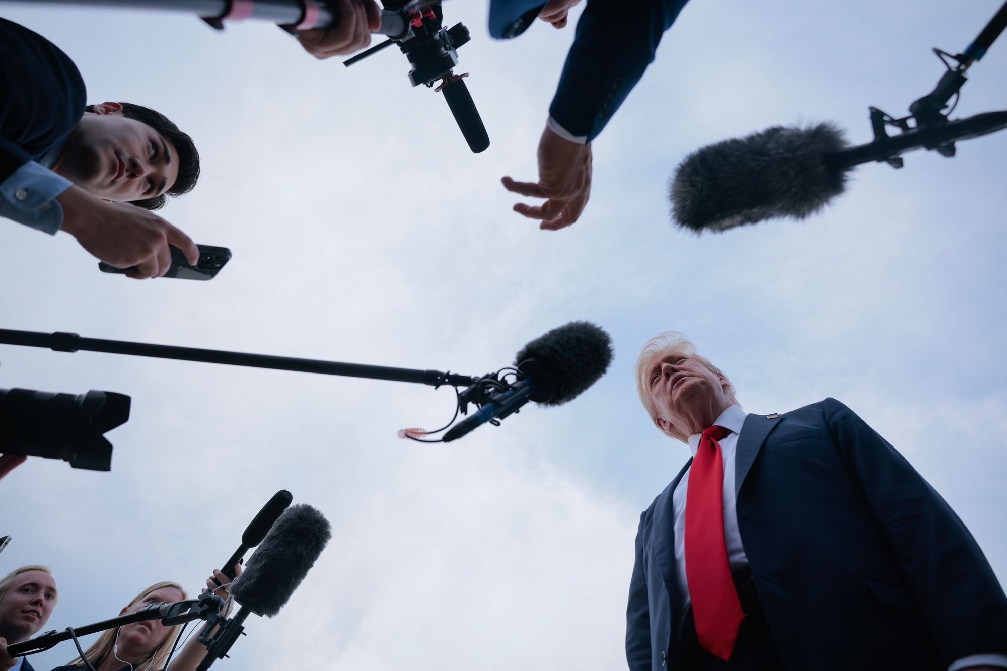 President Donald Trump appears in a photo on the bottom right, with the photographer's angle shooting up from the ground in Trump's direction. Reporters hold microphones, which they are pointing at President Trump.  