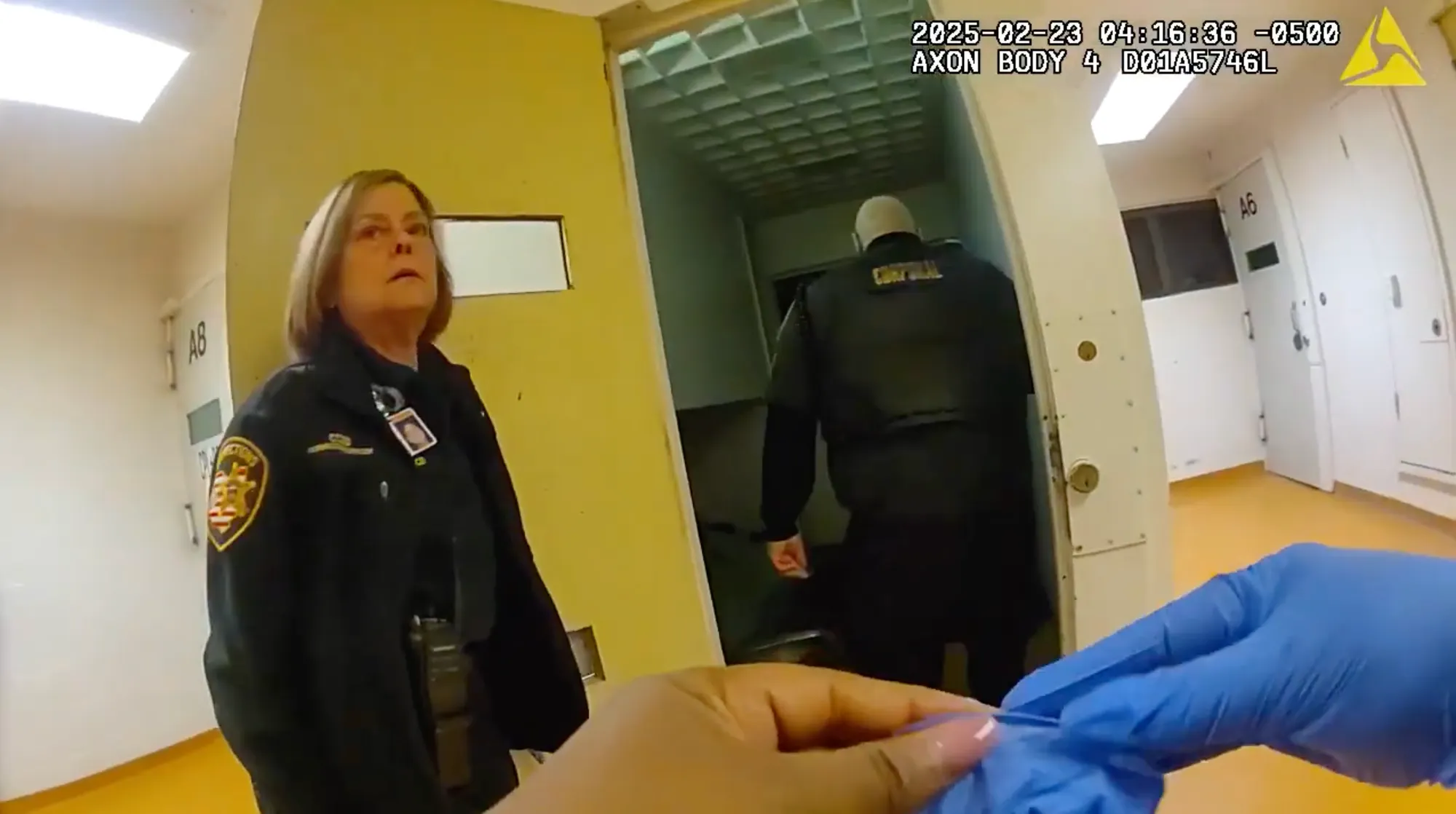 A still from a bodycam shows corrections officers outside Jennifer Wade's cell. Her body is not visible. In the foreground is a person putting on rubber gloves.