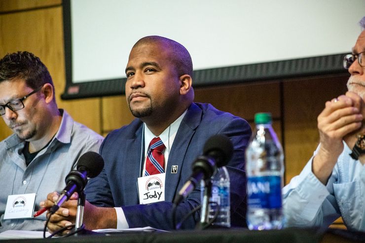 A photo of a bald Black man with a goatee sitting at a podium in front of a microphone. He is wearing a blue suit with a red-and-blue striped tie.