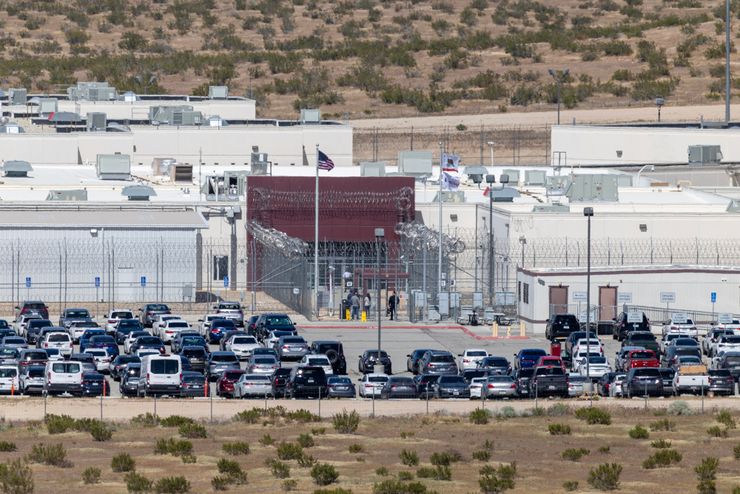 A photo shows the entrance of a detention facility surrounded by barbed wire fencing. Cars are parked in front of the building. 