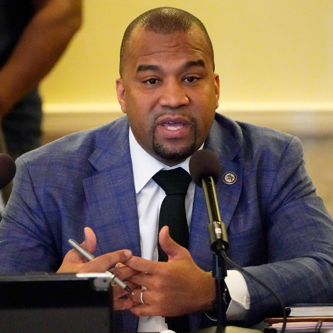 A Black man with a goatee wearing a blue suit jacket speaks in front of a microphone. 