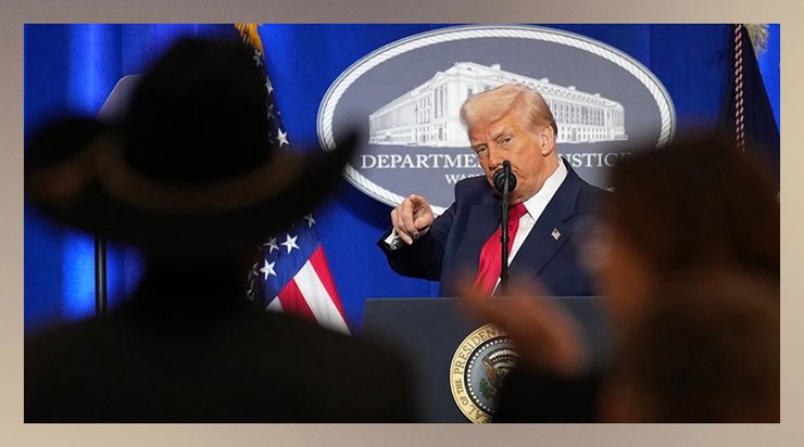President Trump, a White man wearing a navy blue suit jacket, white button-down shirt and red tie, points at members of the audience. In the foreground of the photo, there are silhouettes of audience members, including one person wearing a cowboy hat. Behind Trump is a cobalt blue backdrop and a sign that says “Department of Justice.” 