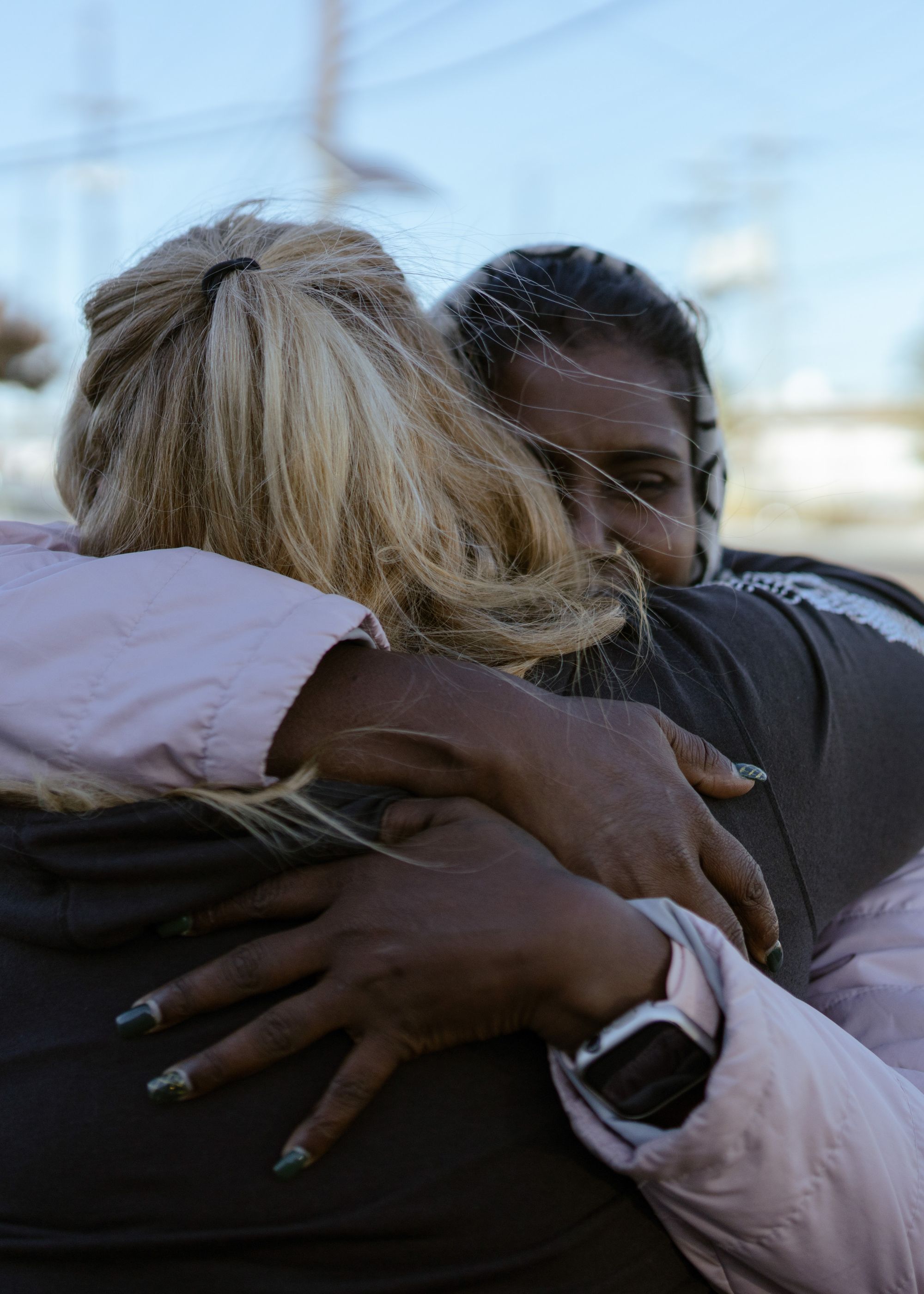 A photo shows a woman, Sandra Hafraoui, with medium-length blonde hair, facing away from the camera as she hugs a woman with dark-toned skin. 