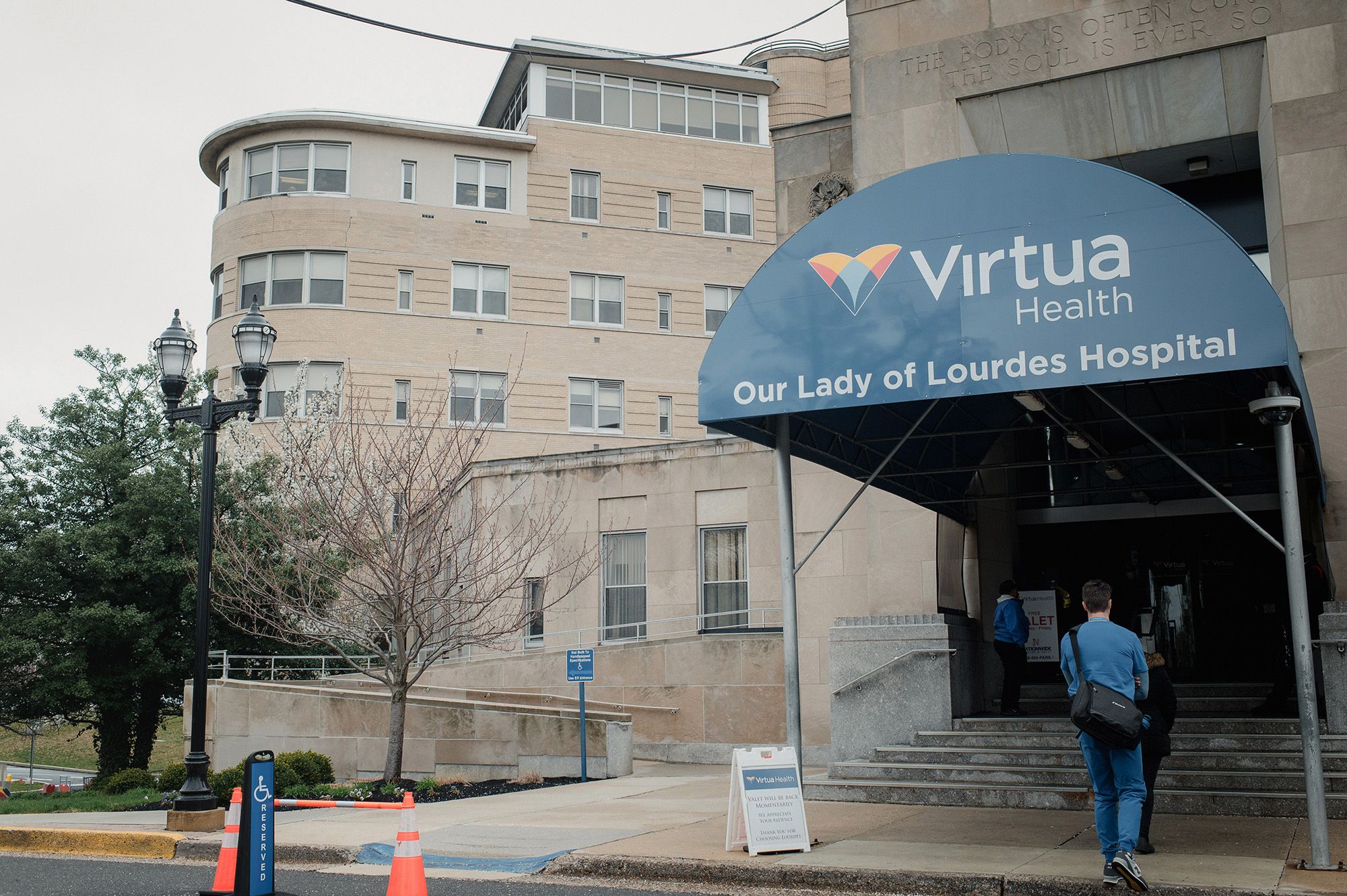 A person wearing a blue uniform and black messenger bag walks toward the entrance of a hospital with a blue sign that reads, "Virtua Health Our Lady of Lourdes Hospital."