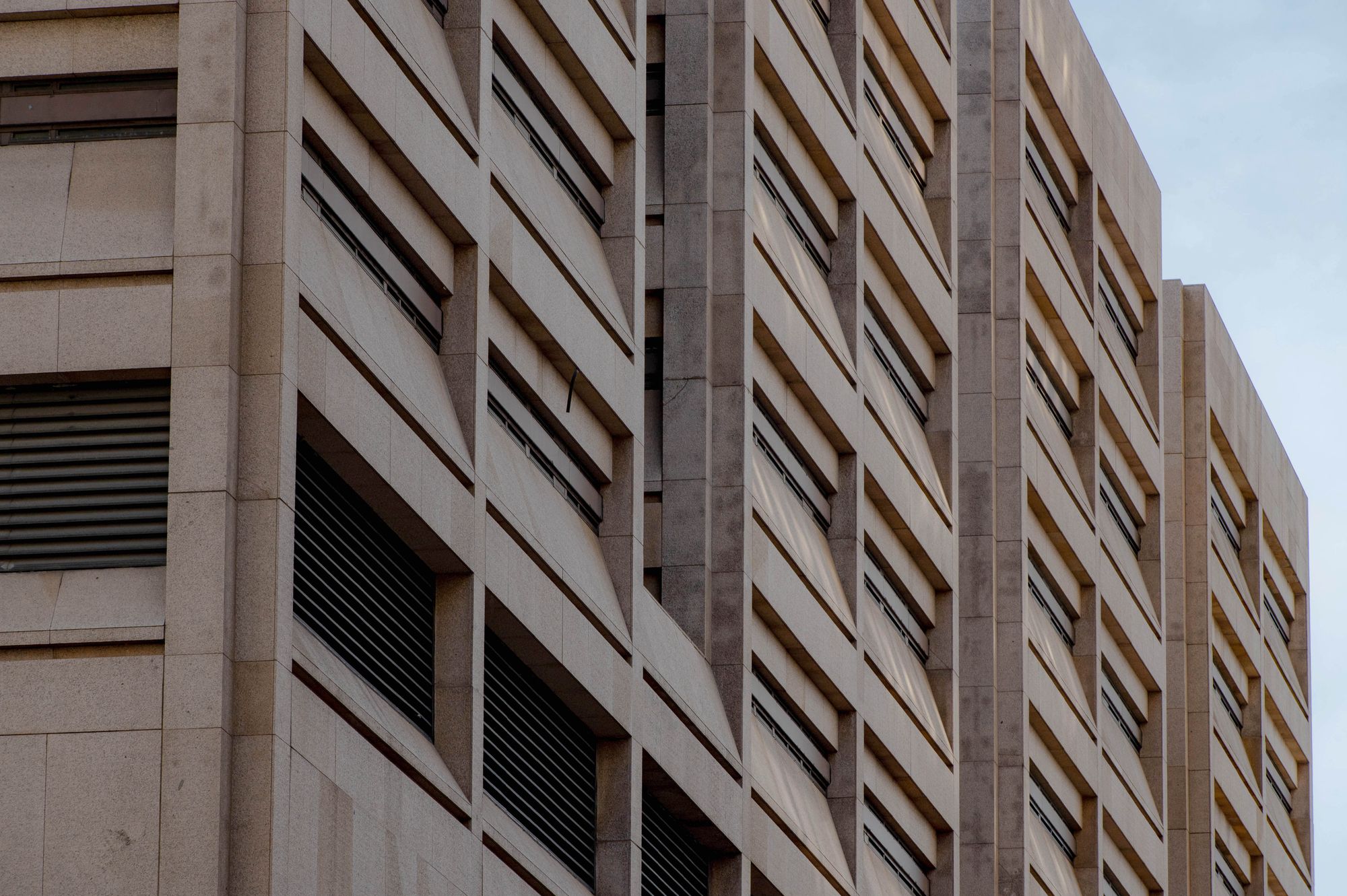 A photo, taken from a low angle, shows the outside of a light brown tower with narrow, horizontal windows. The pale blue sky is visible above the building.