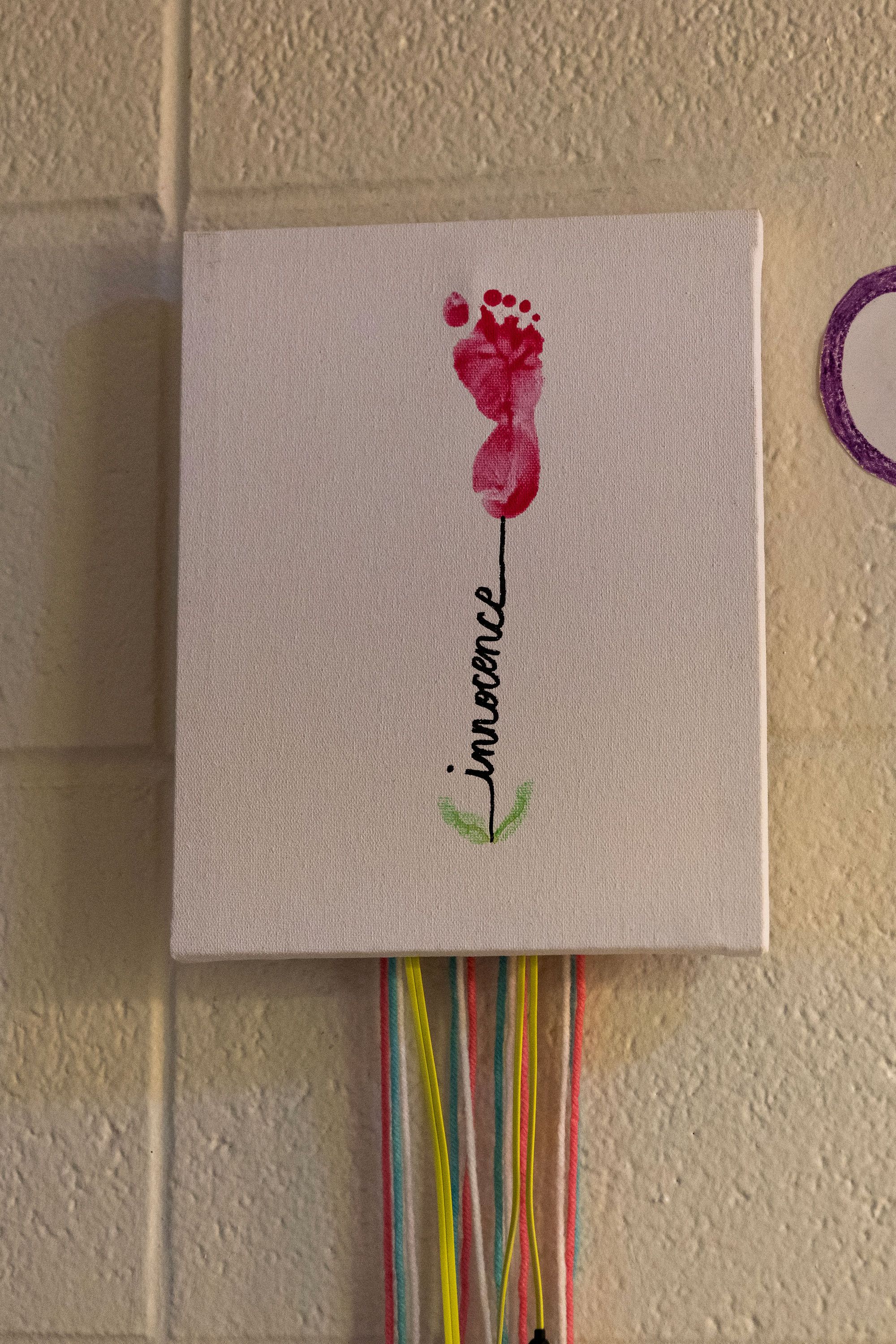 A photo shows a piece of hanging art with a baby’s footprint in pink to represent a flower, and the baby’s name, “Innocence,” representing the stem of the flower.